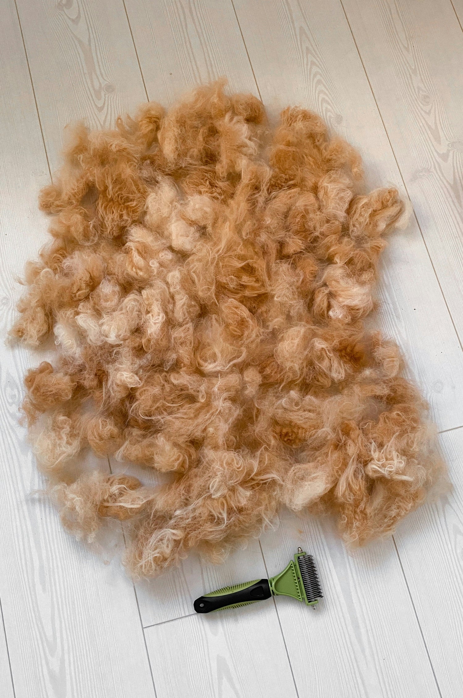 A large pile of golden dog fur next to a green de-shedding brush on a white floor, showing the result of fur removal.